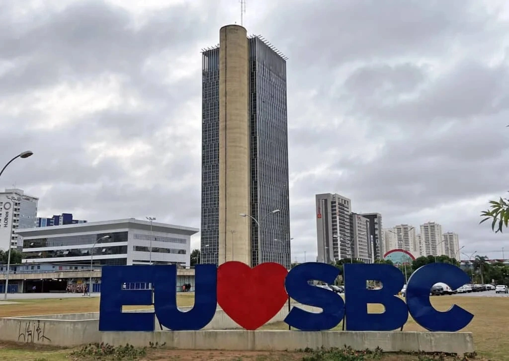 Letreiro “Eu amo SBC” com um coração, em frente a um prédio alto de vidro, em São Bernardo do Campo